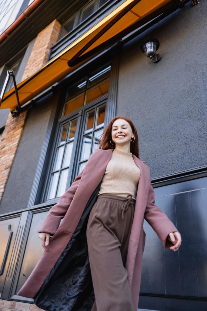 низький кут зору щасливої рудої жінки в стильному пальто, що йде біля сучасної будівлі
  - Фото, зображення