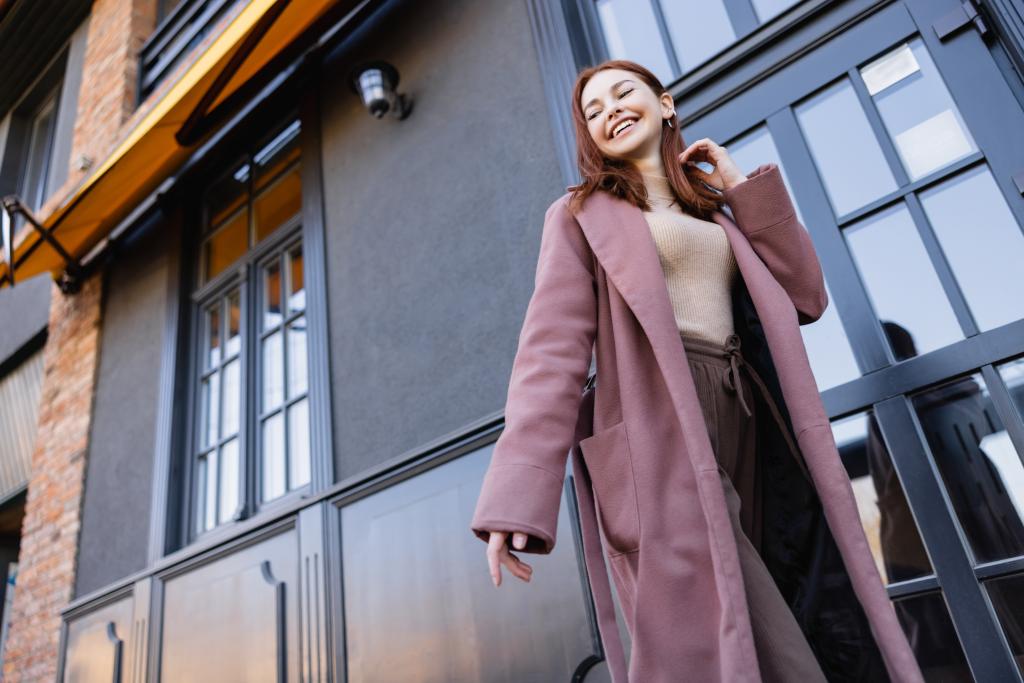 Niedrigwinkel-Ansicht positiver und rothaariger Frau in stylischem Mantel, die in der Nähe moderner Gebäude spaziert  - Foto, Bild