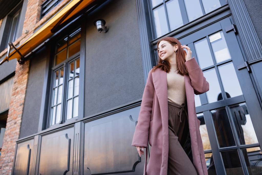 low angle view of happy woman in stylish coat adjusting red hair near modern building - 写真・画像