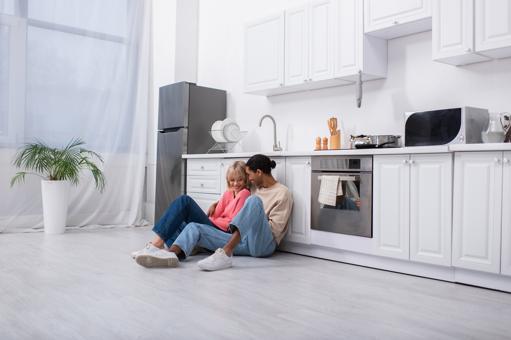 alegre casal multiétnico sentado no chão na cozinha moderna - Foto, Imagem