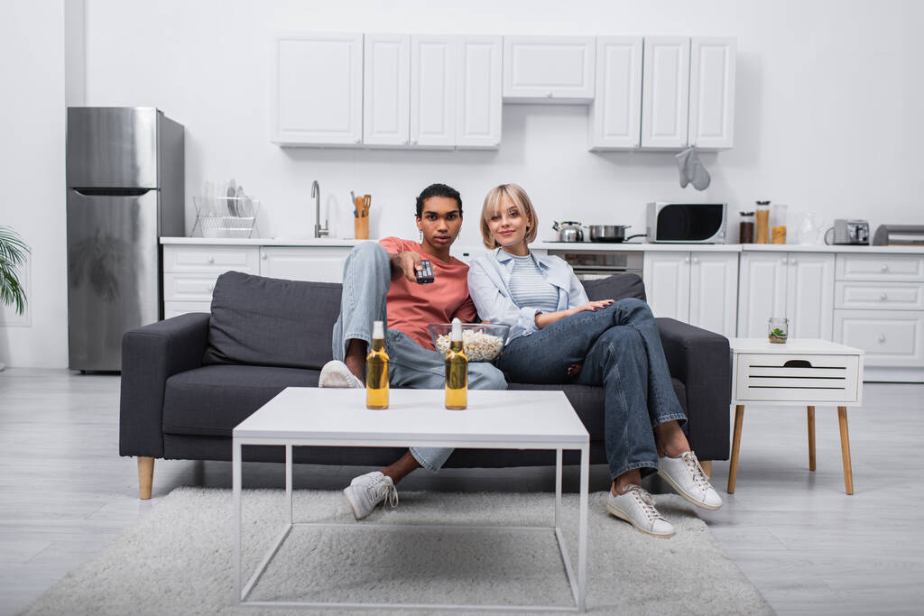 african american man holding remote controller and clicking channels near blonde girlfriend in living room  - Fotografie, Obrázek