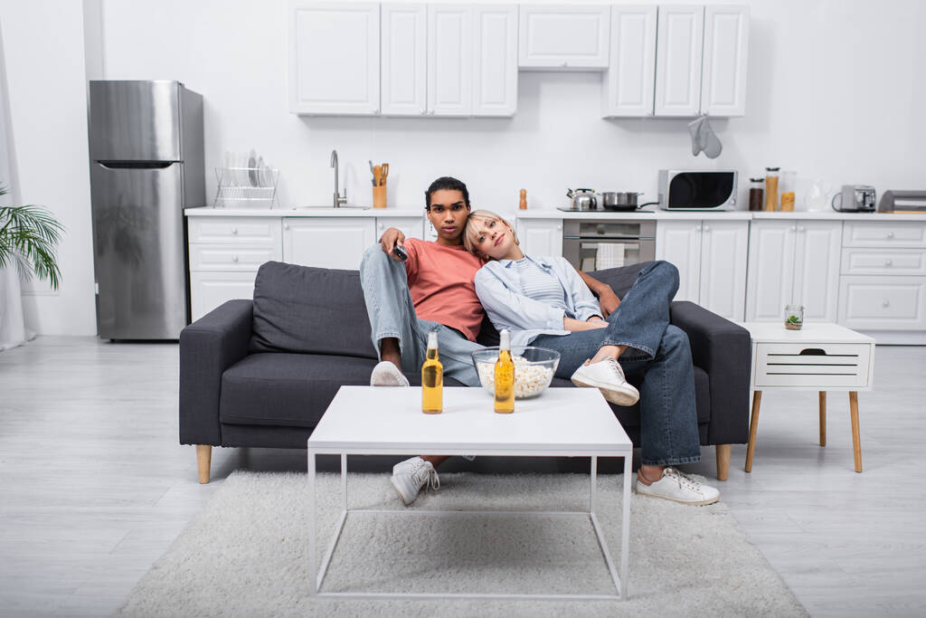 young blonde woman leaning on african american man holding remote controller in living room  - Photo, image