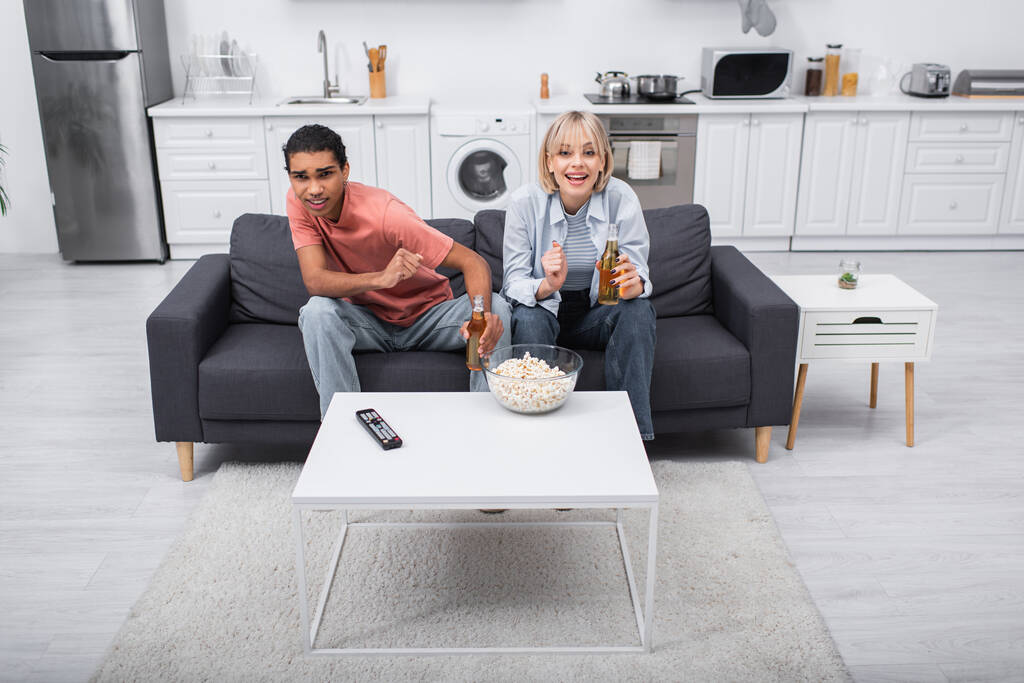 emotiva coppia multietnica tifo e bottiglie di tenuta con birra mentre si guarda partita di sport in soggiorno  - Foto, immagini