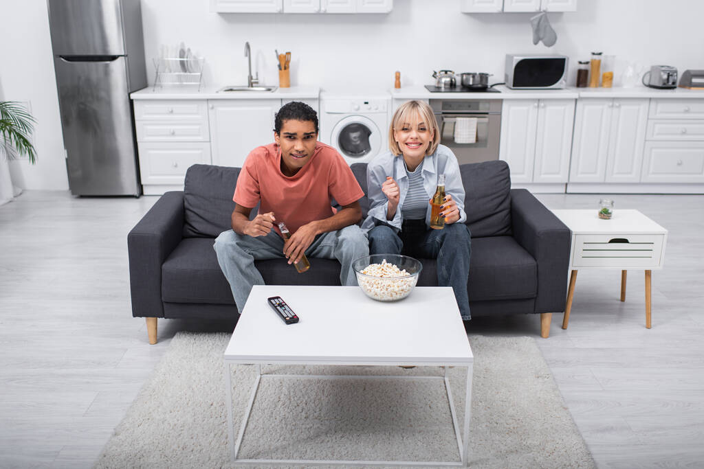 Emotionales gemischtrassiges Paar jubelt und hält Flaschen mit Bier in der Hand, während es sich ein Sportspiel im Wohnzimmer ansieht  - Foto, Bild