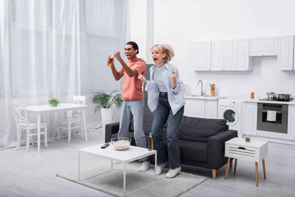excited interracial couple jumping while watching sport match in living room  - Fotoğraf, Görsel