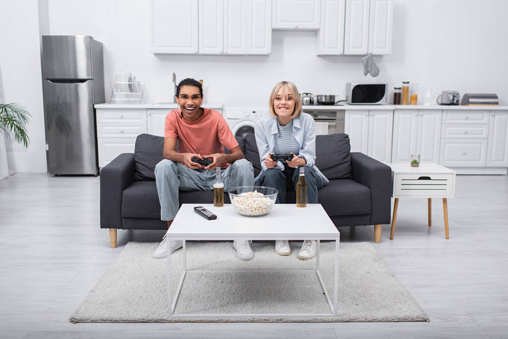 KYIV, UKRAINE - DECEMBER 6, 2021: excited multiethnic couple playing video game in living room  - Foto, afbeelding