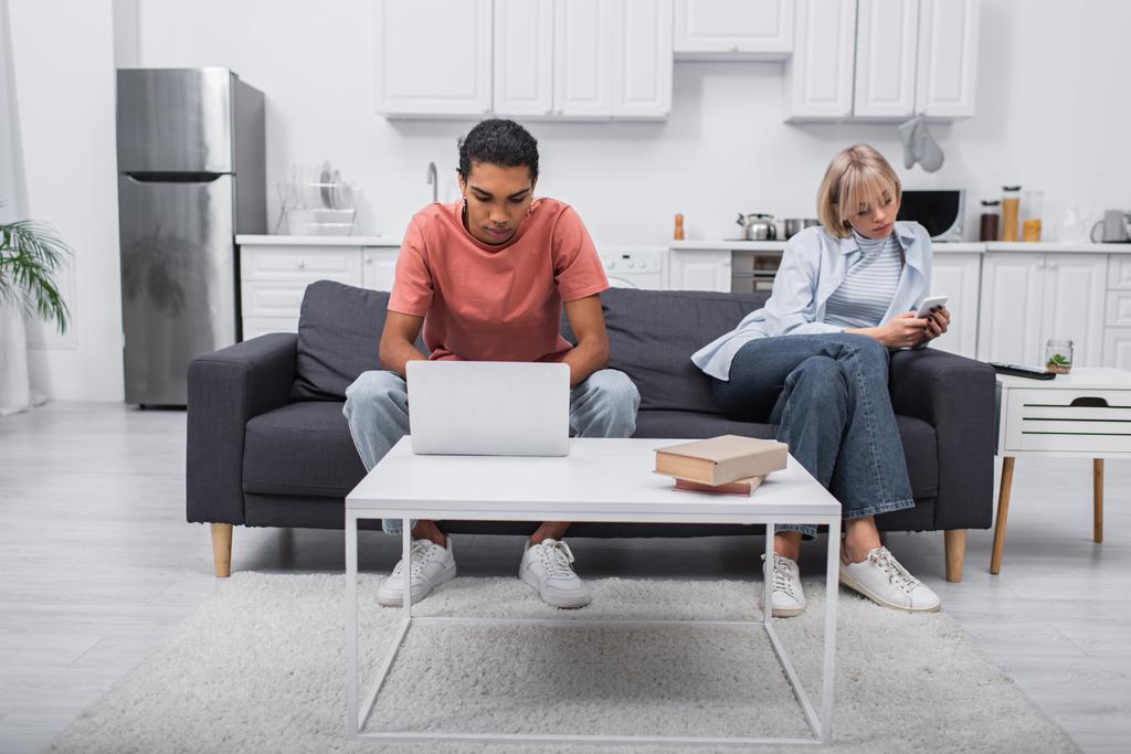 african american man using laptop near girlfriend with smartphone sitting on couch  - Valokuva, kuva