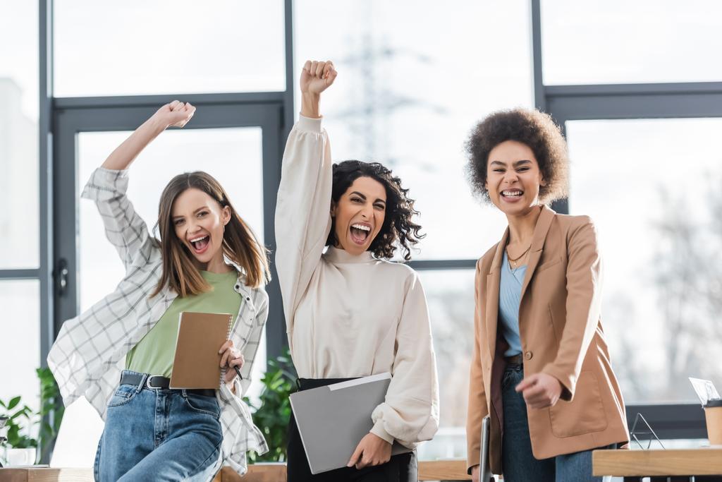 Excited multicultural businesswomen with papers and laptop showing yes gesture in office  - Фото, изображение