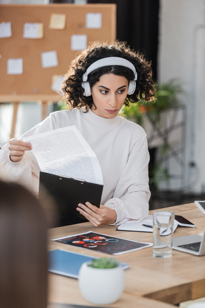 Arabo donna d'affari in cuffie che tengono appunti vicino a dispositivi in ufficio  - Foto, immagini