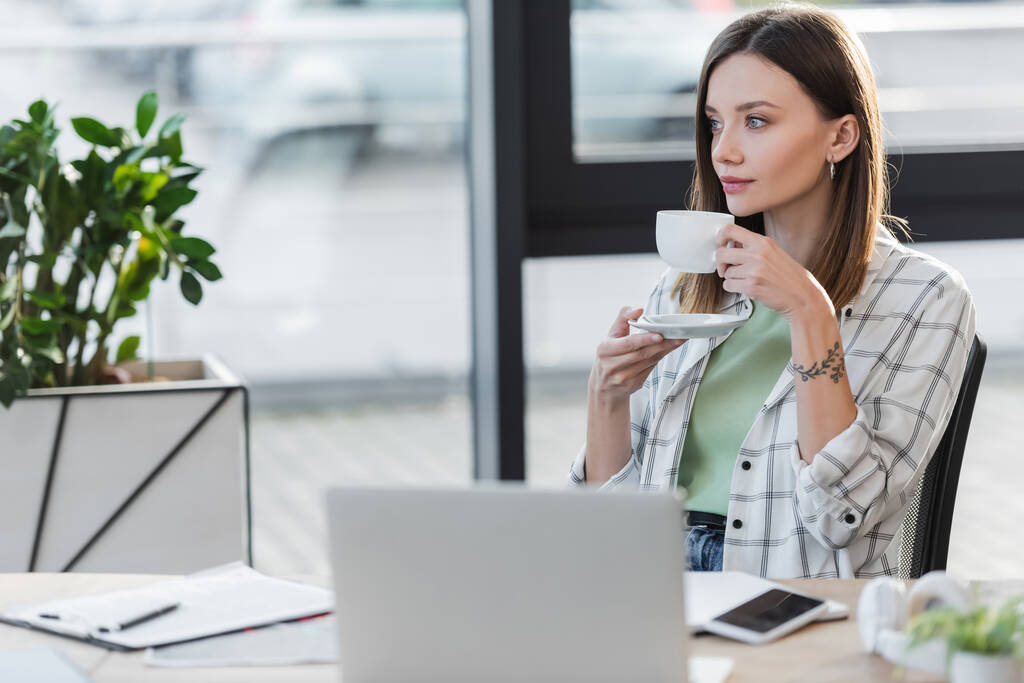 Giovane donna d'affari in possesso di tazza vicino dispositivi sfocati in ufficio  - Foto, immagini