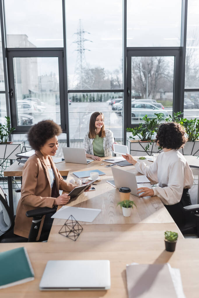 Positieve interraciale zakenvrouwen praten in de buurt van apparaten en papieren op tafel in kantoor  - Foto, afbeelding