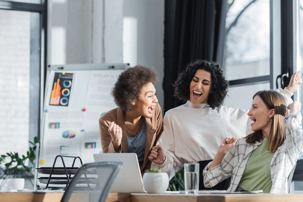 Empresárias multiétnicas excitadas olhando umas para as outras perto do laptop no escritório  - Foto, Imagem