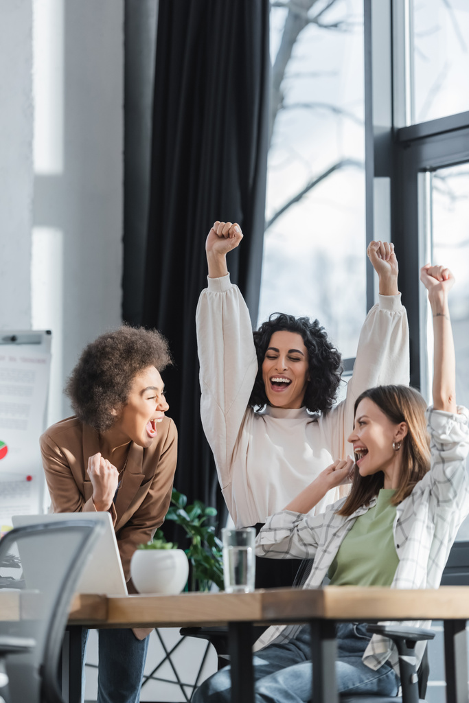 Empresárias multiétnicas entusiasmadas mostrando sim gesto perto de laptop na mesa no escritório  - Foto, Imagem