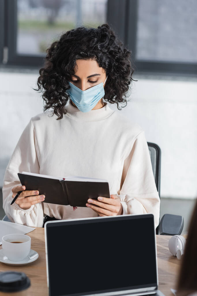 Arabska bizneswoman w masce medycznej trzyma notatnik w pobliżu laptopa i kawy w biurze  - Zdjęcie, obraz
