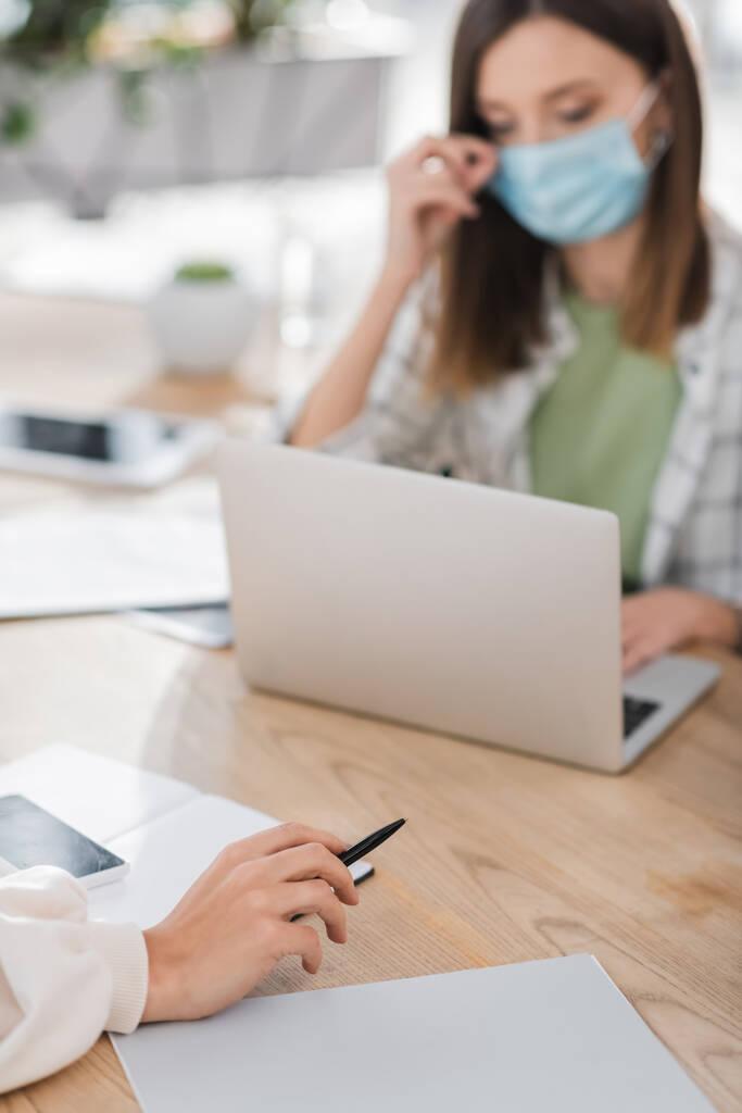 Femme d'affaires tenant un stylo près d'un ordinateur portable, d'un téléphone portable et d'un collègue flou au bureau  - Photo, image