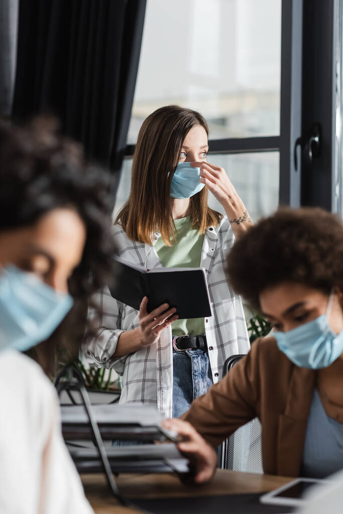 Junge Geschäftsfrau hält Notizbuch in der Hand und trägt medizinische Maske neben verschwommenen multiethnischen Kollegen im Büro  - Foto, Bild