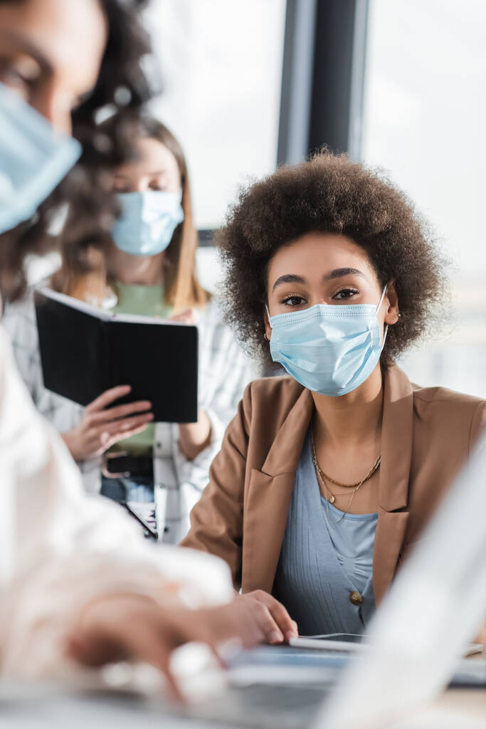 African american businesswoman in medical mask looking at camera near blurred interracial colleagues in office  - Foto, immagini
