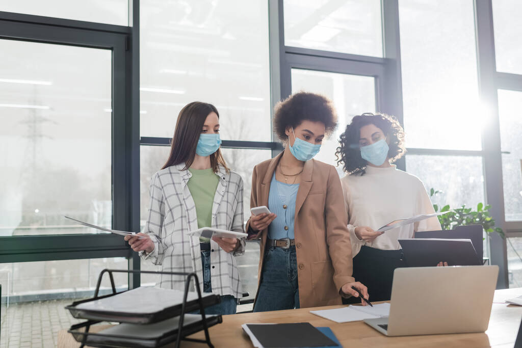 Multiethnic businesswomen in medical masks holding devices and working with papers in office  - Photo, Image