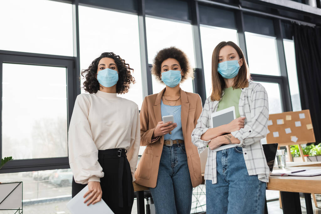 Interracial businesswomen in medical masks holding devices and papers in office  - Photo, Image
