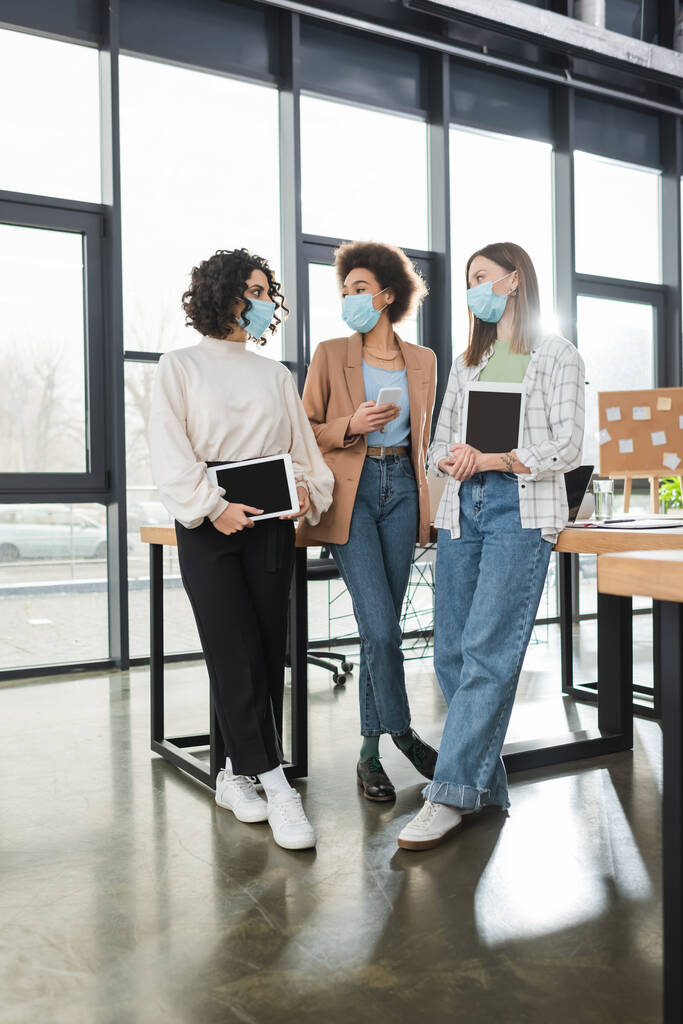 Multiethnische Geschäftsfrauen in medizinischen Masken halten Geräte in der Hand, während sie im Büro reden  - Foto, Bild