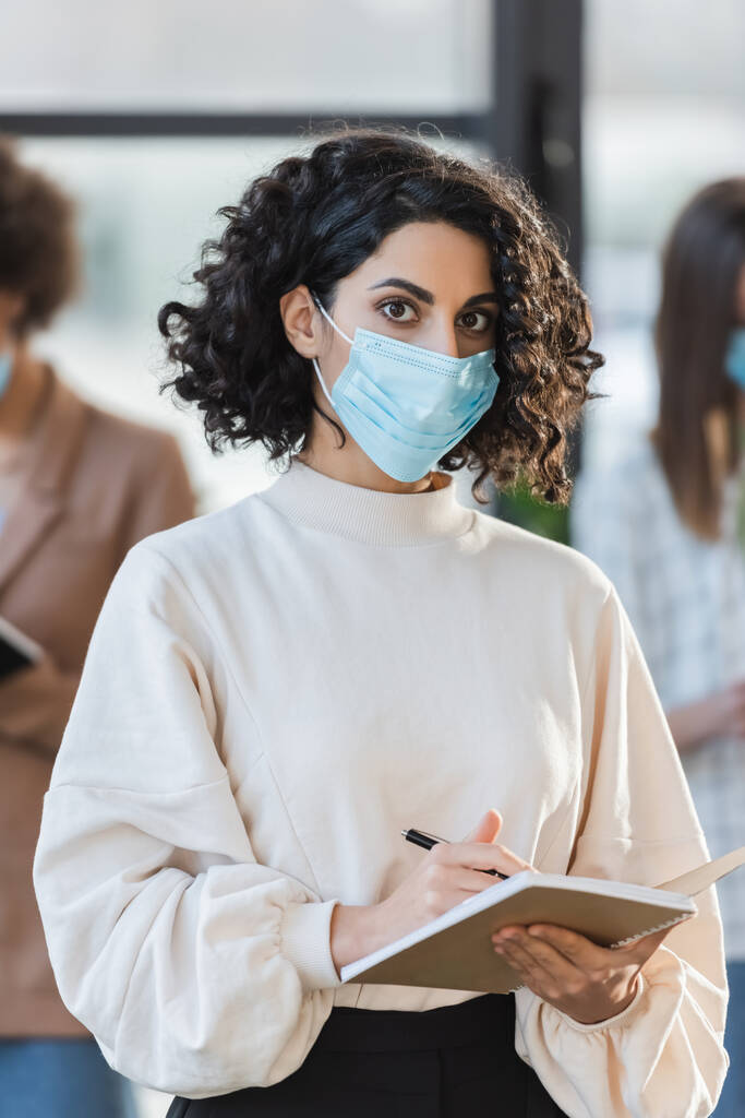 Arabian businesswoman in protective mask holding pen and notebook while looking at camera in office  - Photo, image