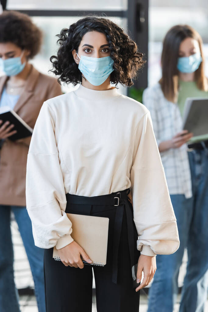 Muslimská podnikatelka v lékařské masce drží notebook a dívá se do kamery v kanceláři  - Fotografie, Obrázek
