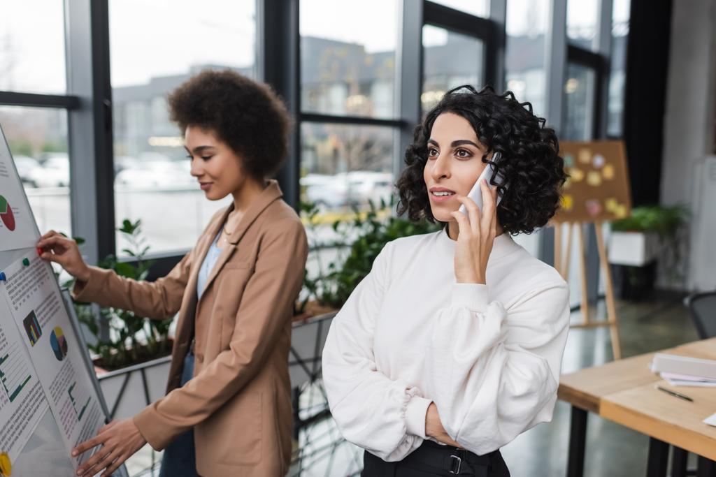 Молодая арабская бизнесвумен разговаривает на смартфоне рядом с размытым африканским коллегой и флип-чартом в офисе - Фото, изображение