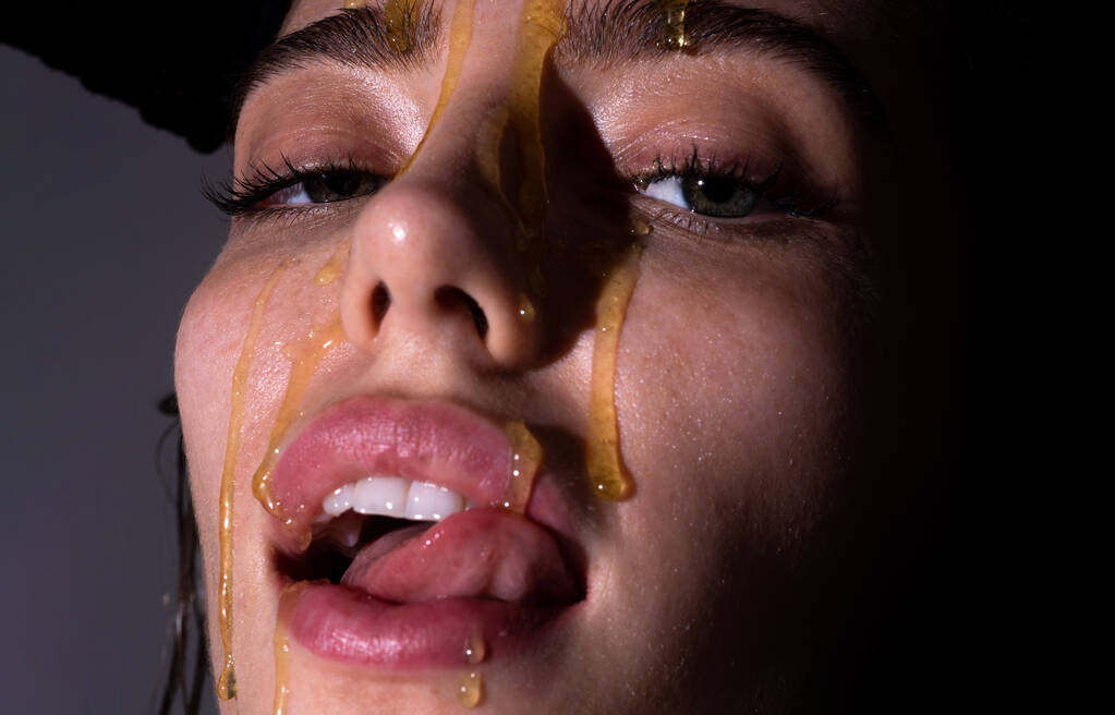 Sensual young woman face with honey drop close up. Sexy model with honey drip on face. Beauty woman face portrait in studio. Beautiful girl tongue licking honey lips. - Photo, Image