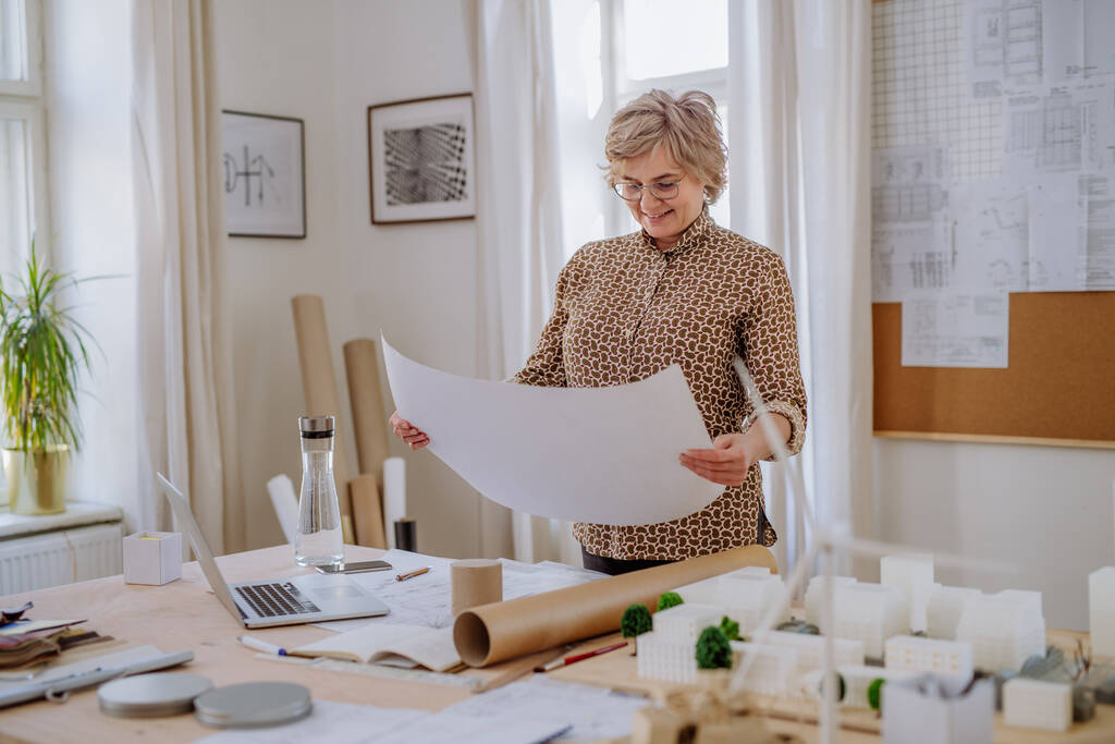 Senior donna architetto con modello di case guardando cianografie in ufficio. - Foto, immagini