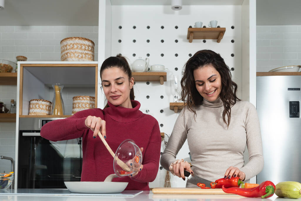 Two young happy college student roommates or business women cooking food together at their apartment. Females having fun together preparing meal in their modern kitchen at cozy home. - Photo, Image