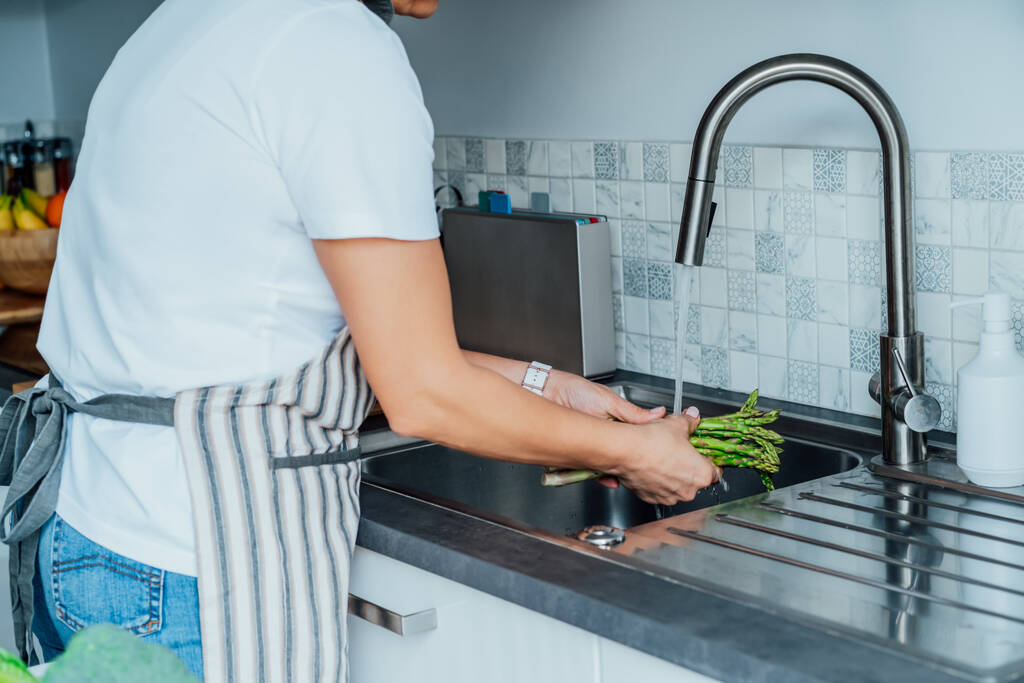 Donna che lava asparagi freschi sotto l'acqua corrente sopra lavandino sulla sua cucina. Preparare gli ingredienti alimentari per il pranzo. Cucinare cibo sano. Ricette con verdure, dieta equilibrata. - Foto, immagini
