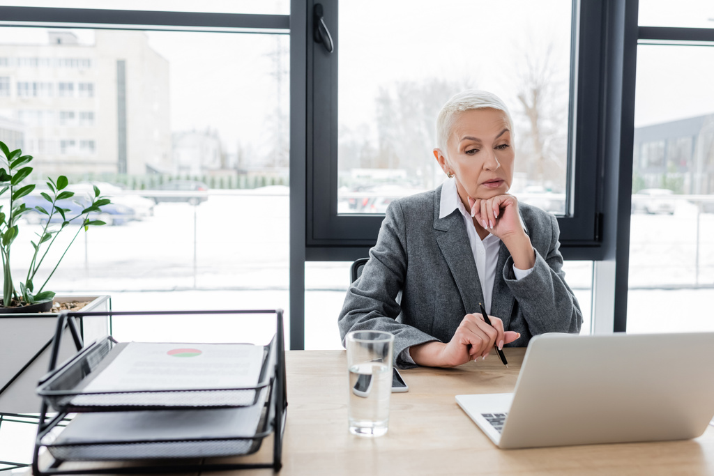 thoughtful businesswoman looking at laptop near glass of water on desk - Фото, изображение