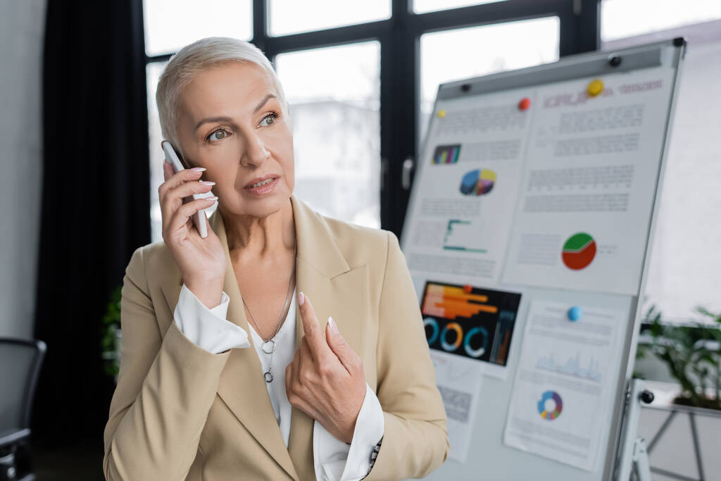 stylish businesswoman talking on mobile phone near flip chart on blurred background - Foto, Bild