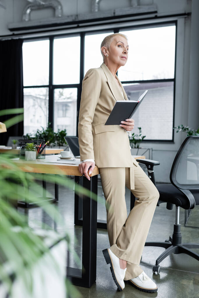 full length view of stylish banker standing with notebook near desk and looking away - Photo, image