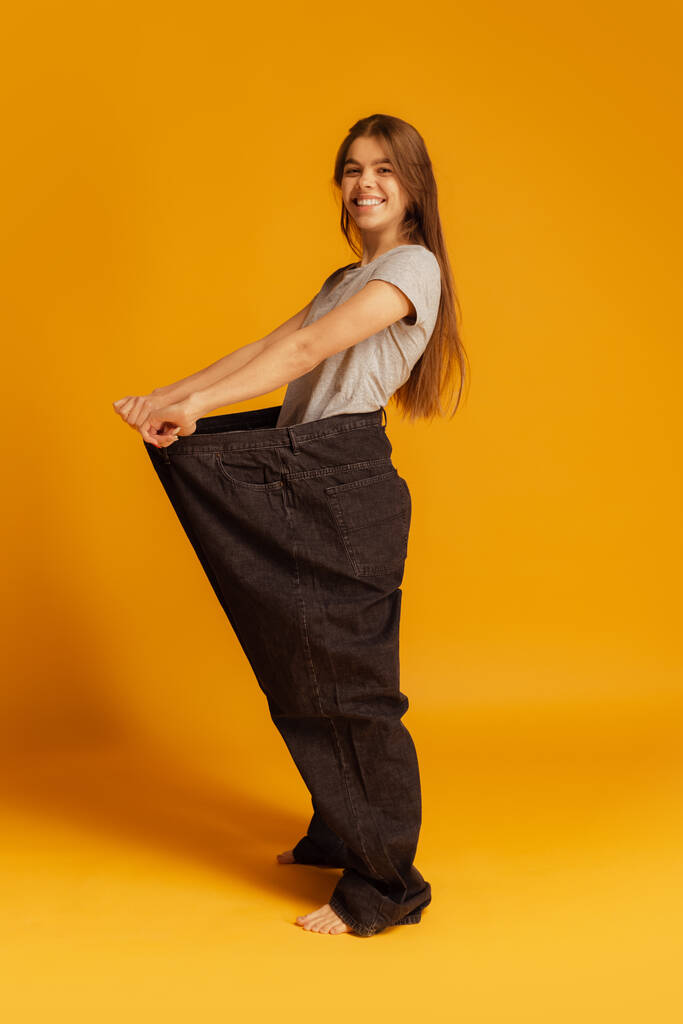 Vista panorámica. Retrato de una joven mujer feliz de pie en pantalones negros gigantes aislados sobre fondo amarillo. Concepto de pérdida de peso - Foto, Imagen