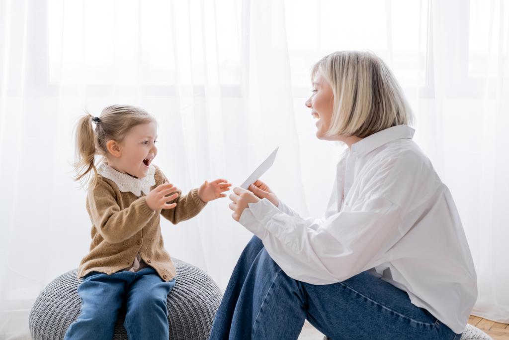 cheerful blonde woman holding card near amazed daughter while sitting on poufs at home - Photo, Image