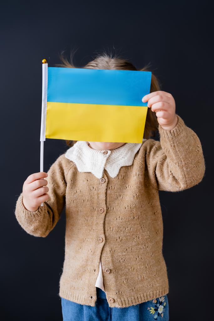 patriotic girl obscuring face with small ukrainian flag isolated on black - Photo, Image