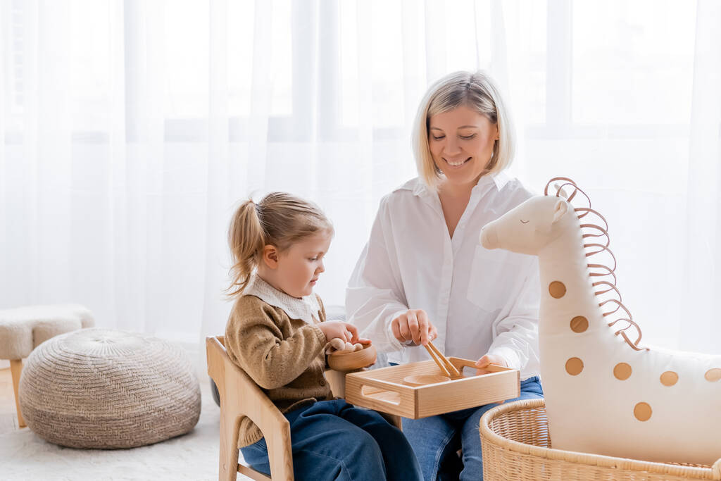 feliz madre e hija jugando con cuencos de madera y palos cerca de caballo de juguete en casa - Foto, Imagen