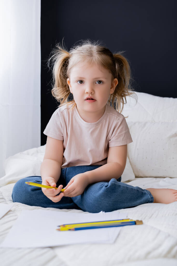 маленькая девочка сидит на кровати рядом с голубыми и желтыми карандашами и смотрит в камеру - Фото, изображение