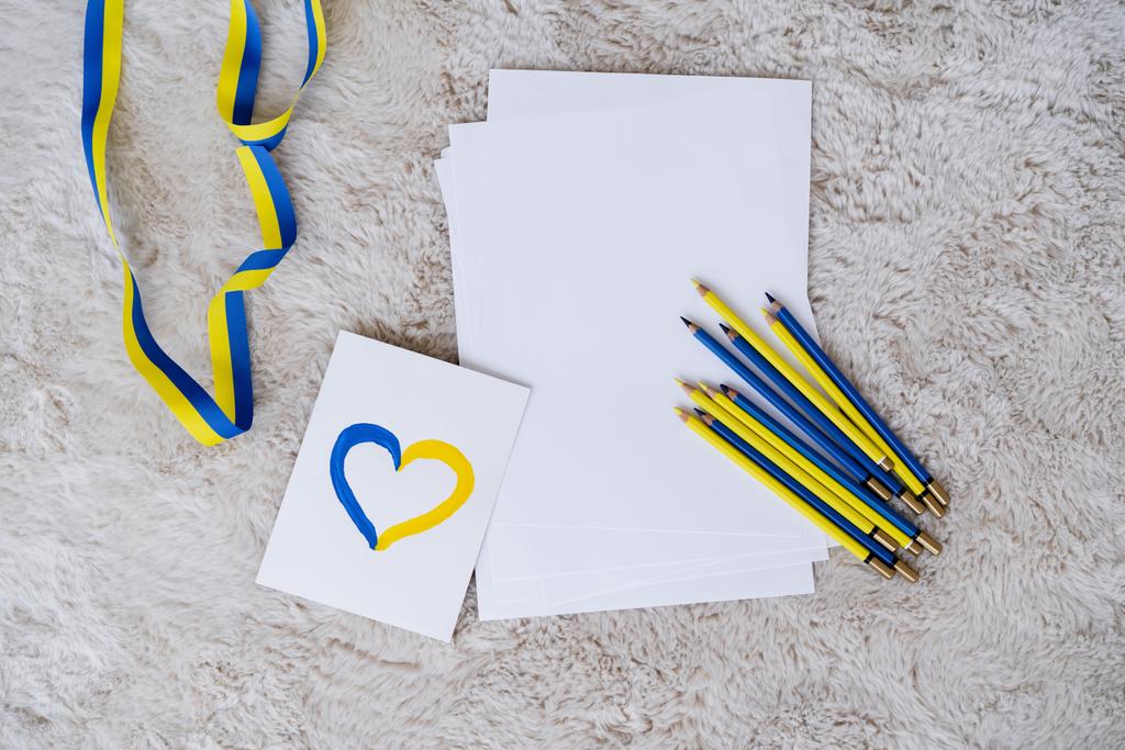 felső nézet színes ceruzák, szalag és kártya kék és sárga szív szürke bolyhos szőnyeg - Fotó, kép