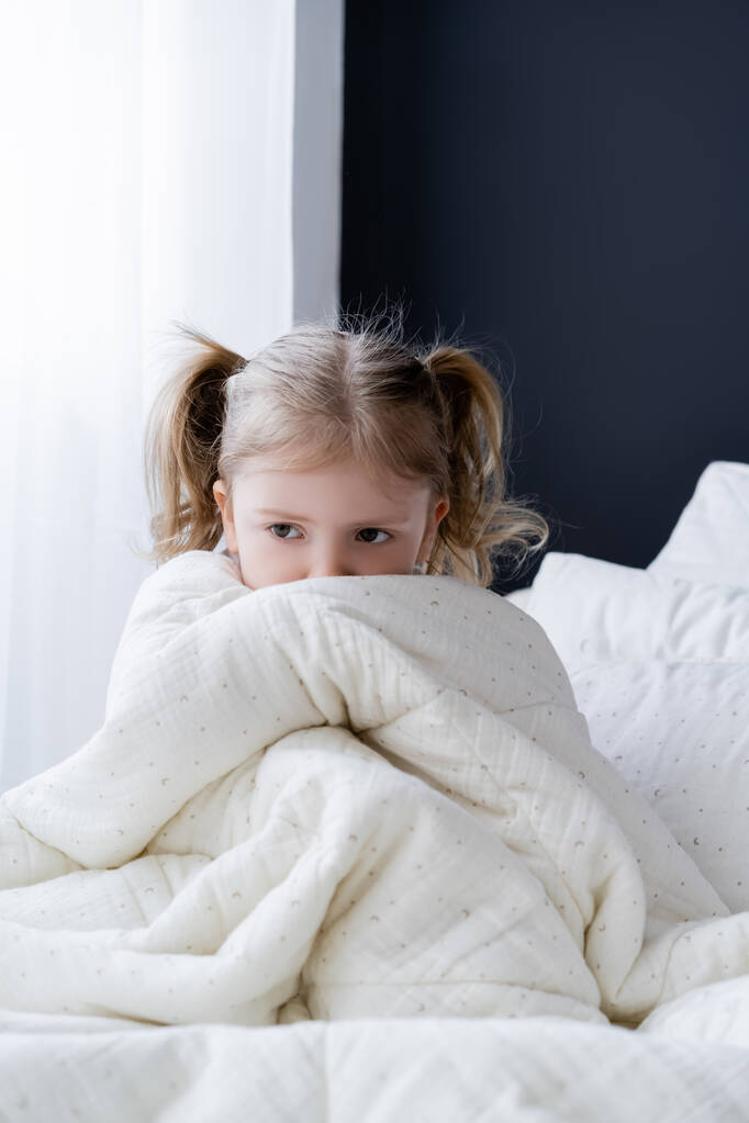 verängstigtes Mädchen sitzt auf Bett und verdeckt Gesicht mit weißer Decke - Foto, Bild