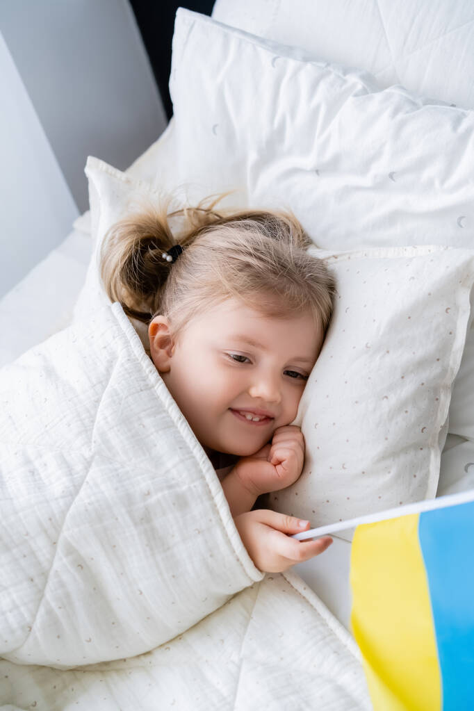 άποψη υψηλής γωνίας του χαμογελαστού πατριωτικό κορίτσι που βρίσκεται στο κρεβάτι με μικρή ουκρανική σημαία - Φωτογραφία, εικόνα