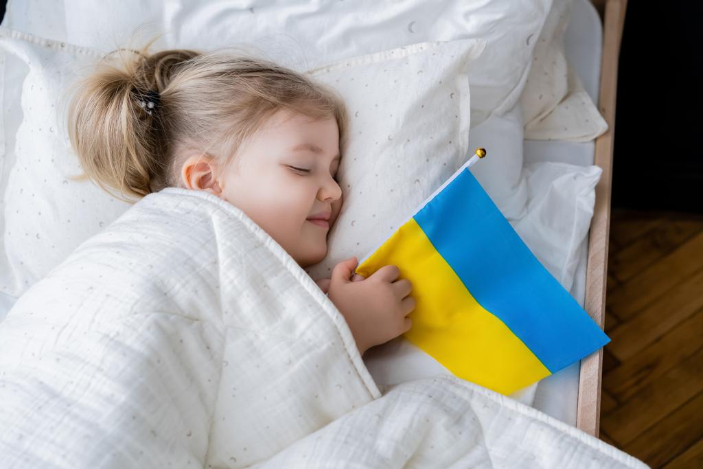 vista superior de la chica sonriente durmiendo en la cama con pequeña bandera ucraniana - Foto, Imagen