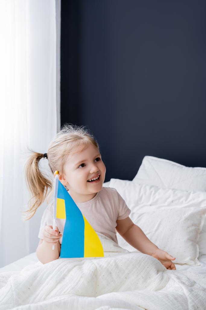 alegre chica sosteniendo pequeña bandera ucraniana mientras está sentado en la cama - Foto, Imagen