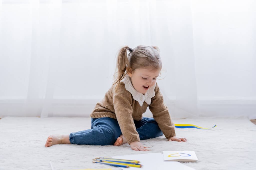menina animado sentado no chão perto de lápis de cor e cartão com coração azul e amarelo - Foto, Imagem