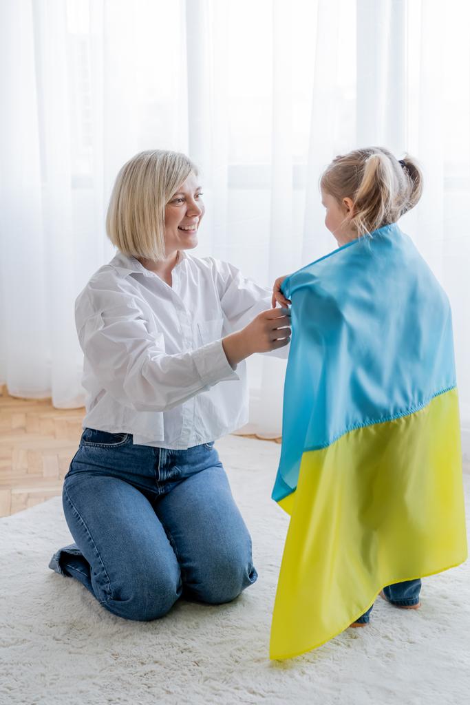 sonriente mujer rubia cubriendo a su hijita con bandera ucraniana en casa - Foto, imagen