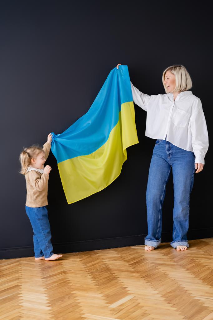 full length view of excited mom and daughter holding ukrainian flag near black wall - Photo, Image