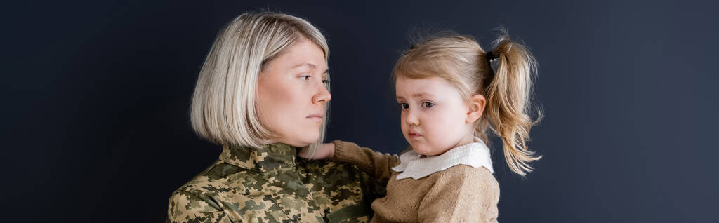 blond vrouw in camouflage kijken naar boos dochter geïsoleerd op zwart, banner - Foto, afbeelding