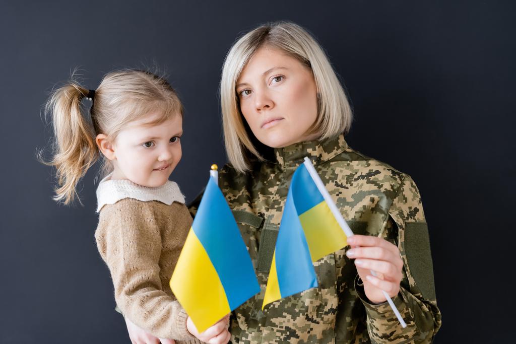 mujer militar con hija pequeña sosteniendo banderas ucranianas pequeñas aisladas en negro - Foto, imagen