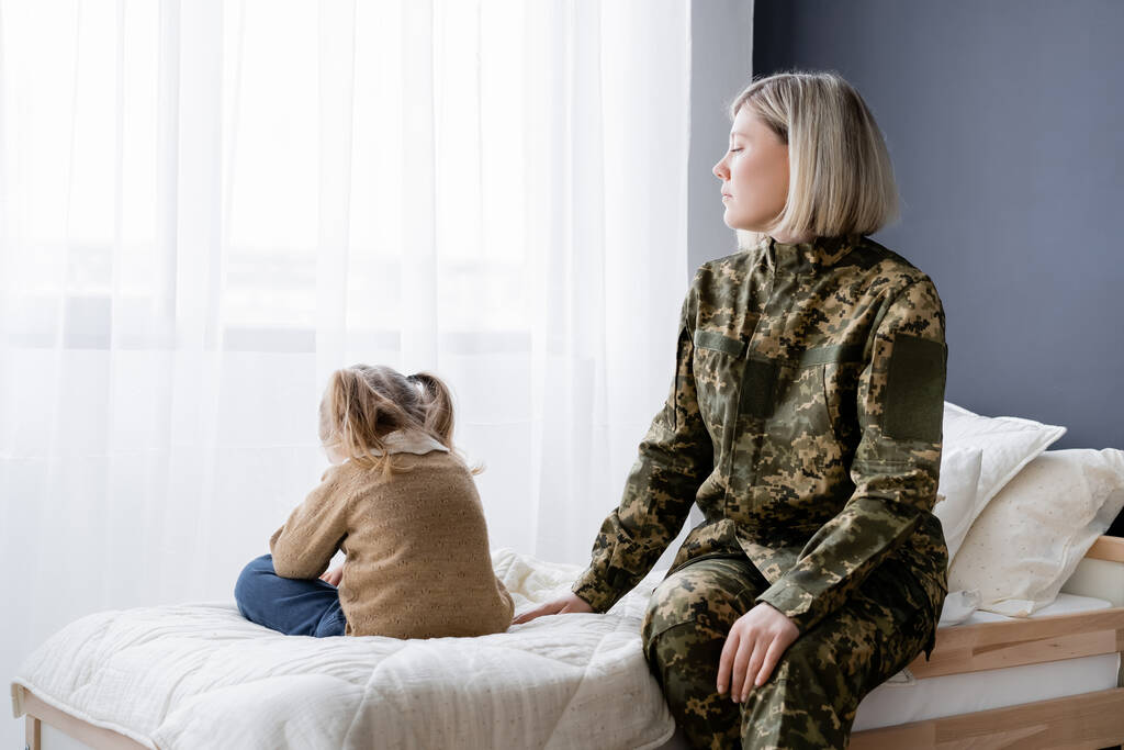 γυναίκα με στρατιωτική στολή κάθεται στο κρεβάτι κοντά προσβεβλημένη κόρη - Φωτογραφία, εικόνα